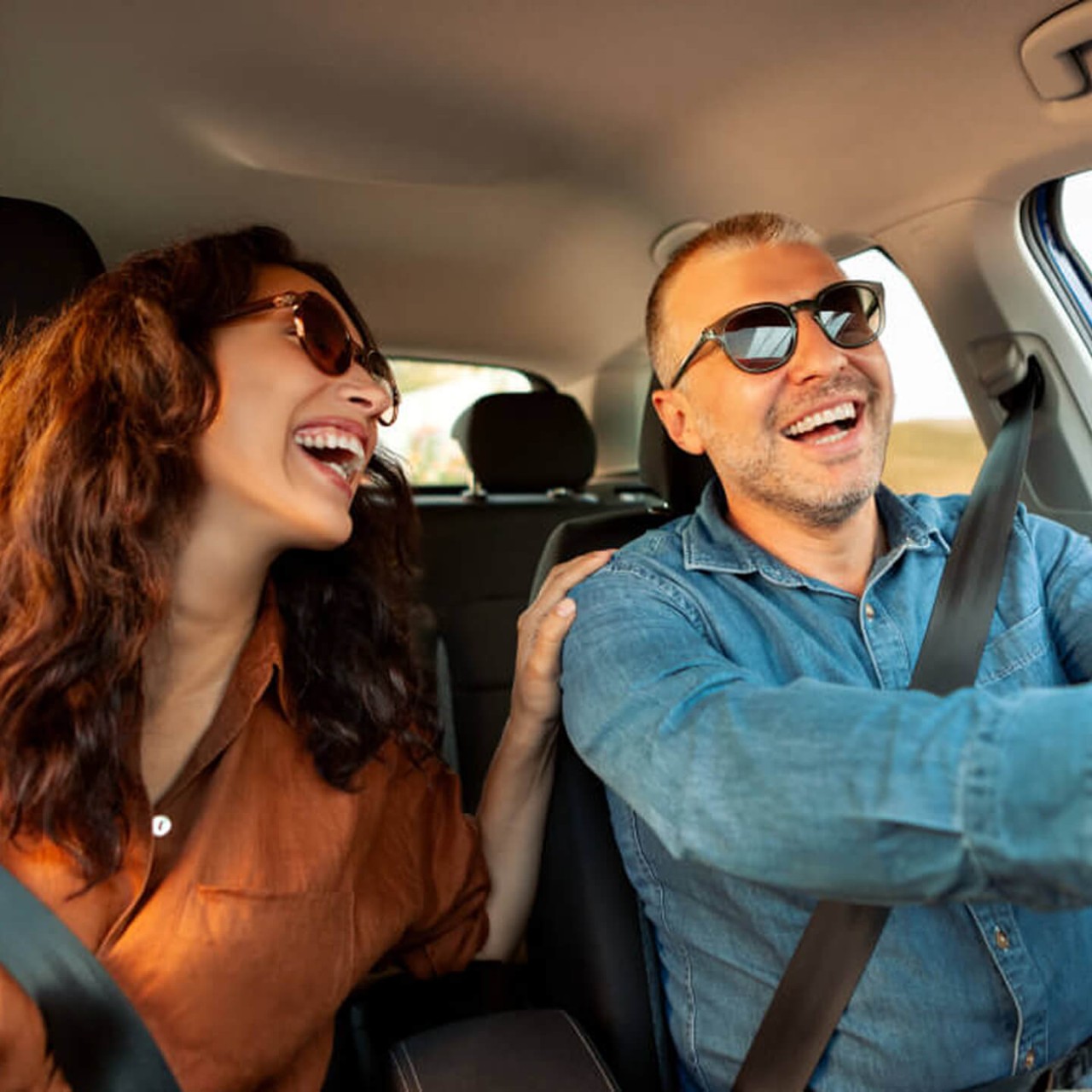 A happy couple enjoys a drive with their sunglasses on. The man smiles, looking at the road while the woman smiles back with her hand on his shoulder.